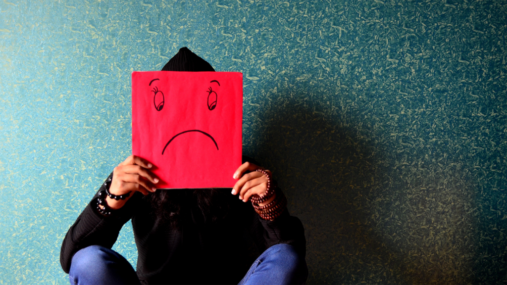 person sitting and holding a red piece of paper with a sad face drawn on it in front of their face