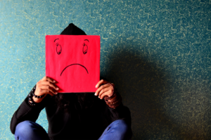 person sitting and holding a red piece of paper with a sad face drawn on it in front of their face