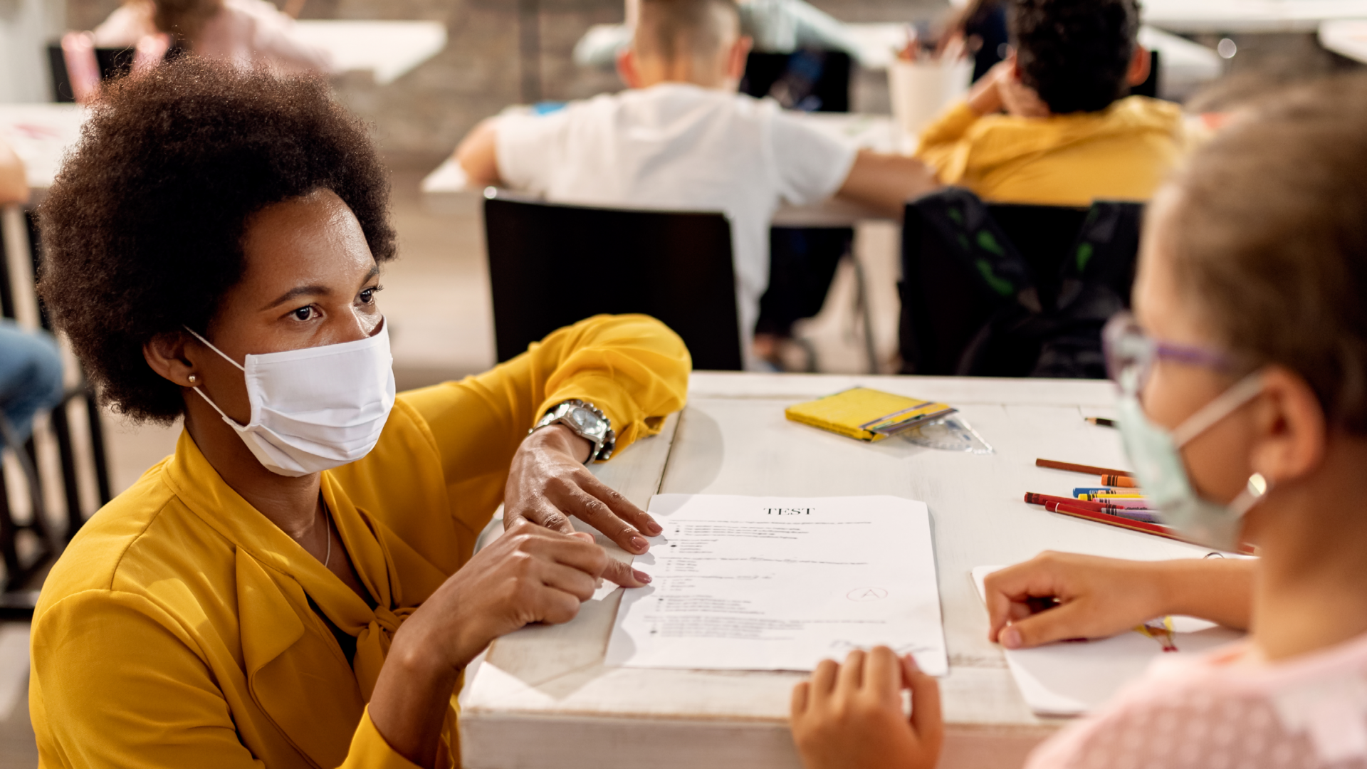 teacher in mask kneeling and speaking to a young girl student with mask at desk