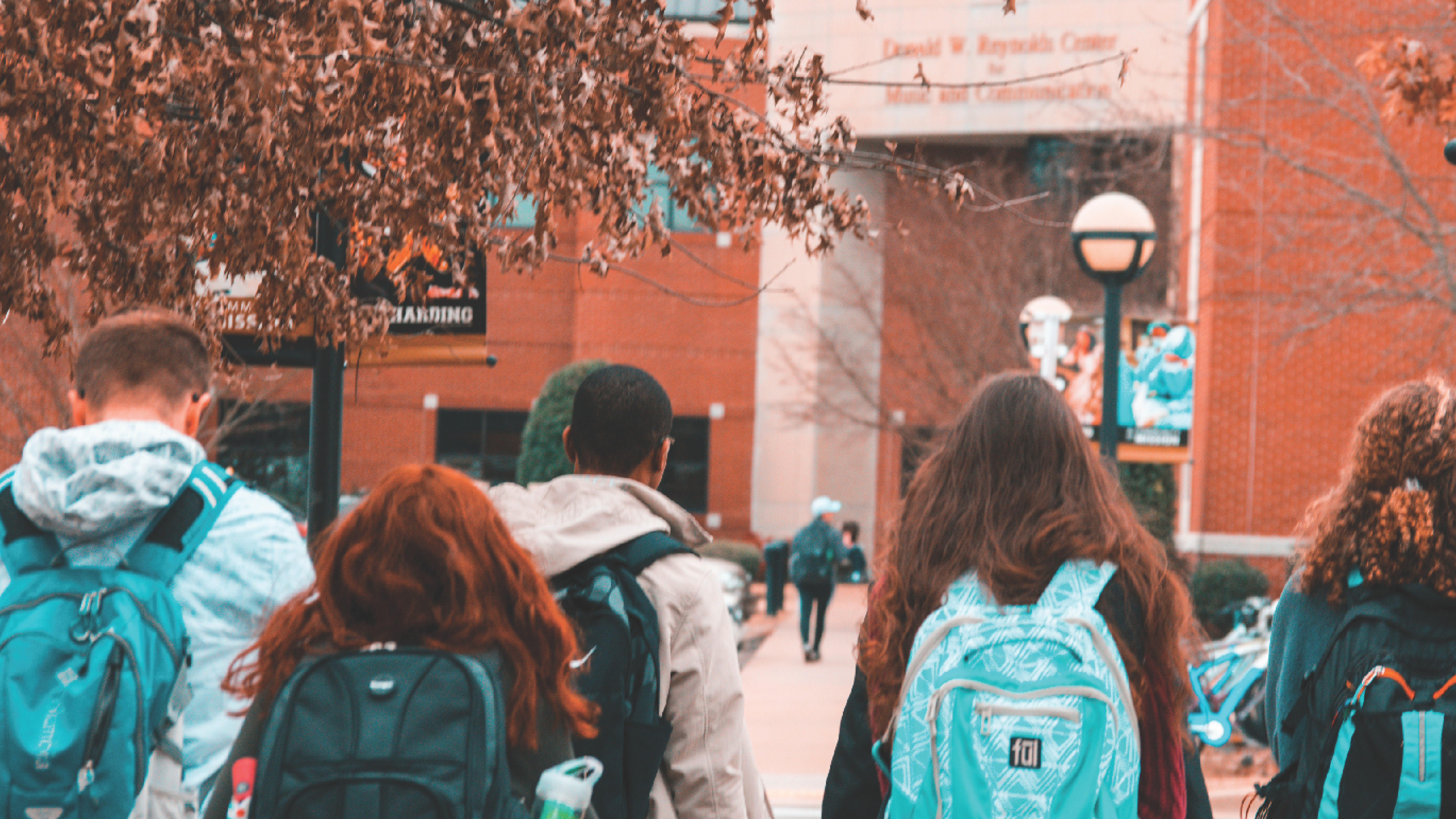 five young adult students with backpacks walking toward school building seen from behind