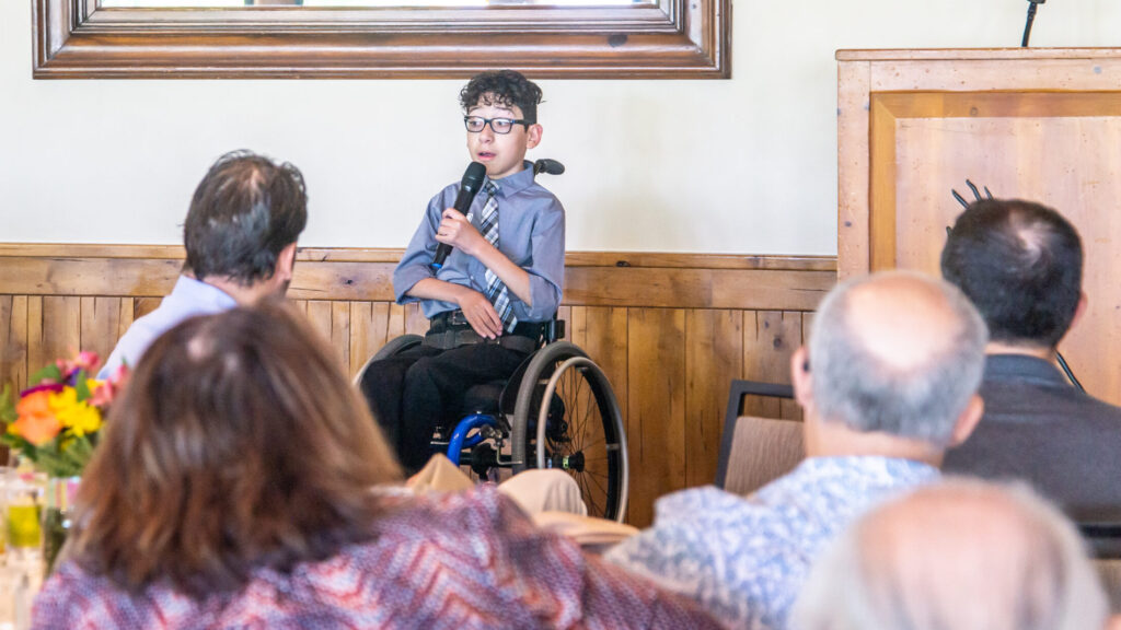 Dark haired teen in wheelchair holding microphone speaking to a group of people from a stage.