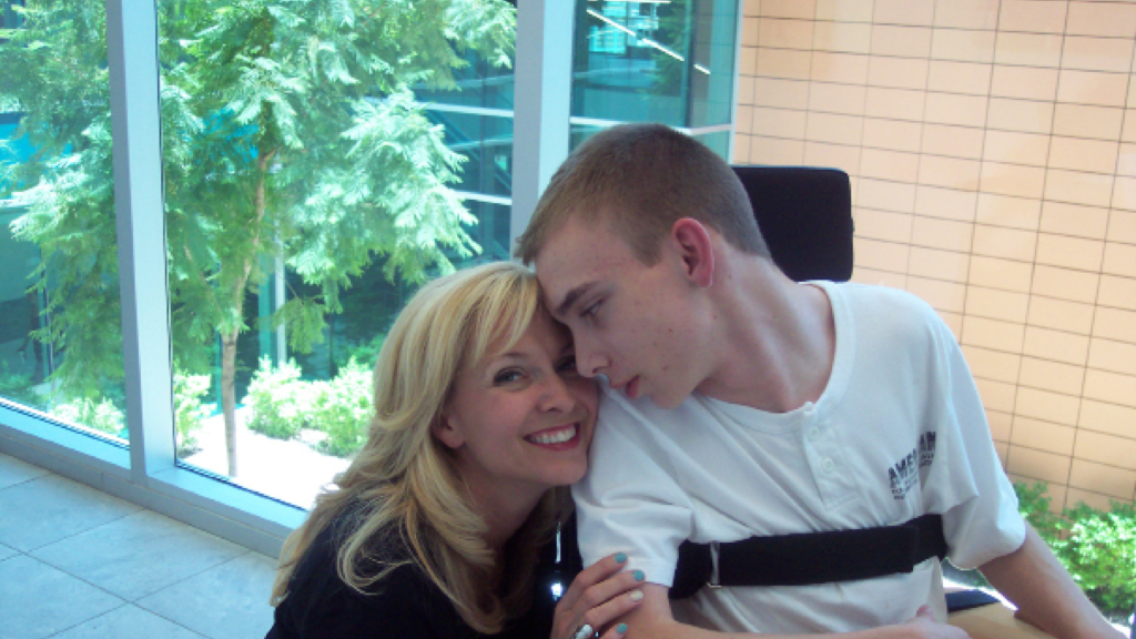 Young man in a wheelchair smiling at his mom who is kneeling next to his chair and smiling at the camera