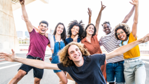 A group of seven young adults smiling at the camera with arms up one front and center with his arms wide open