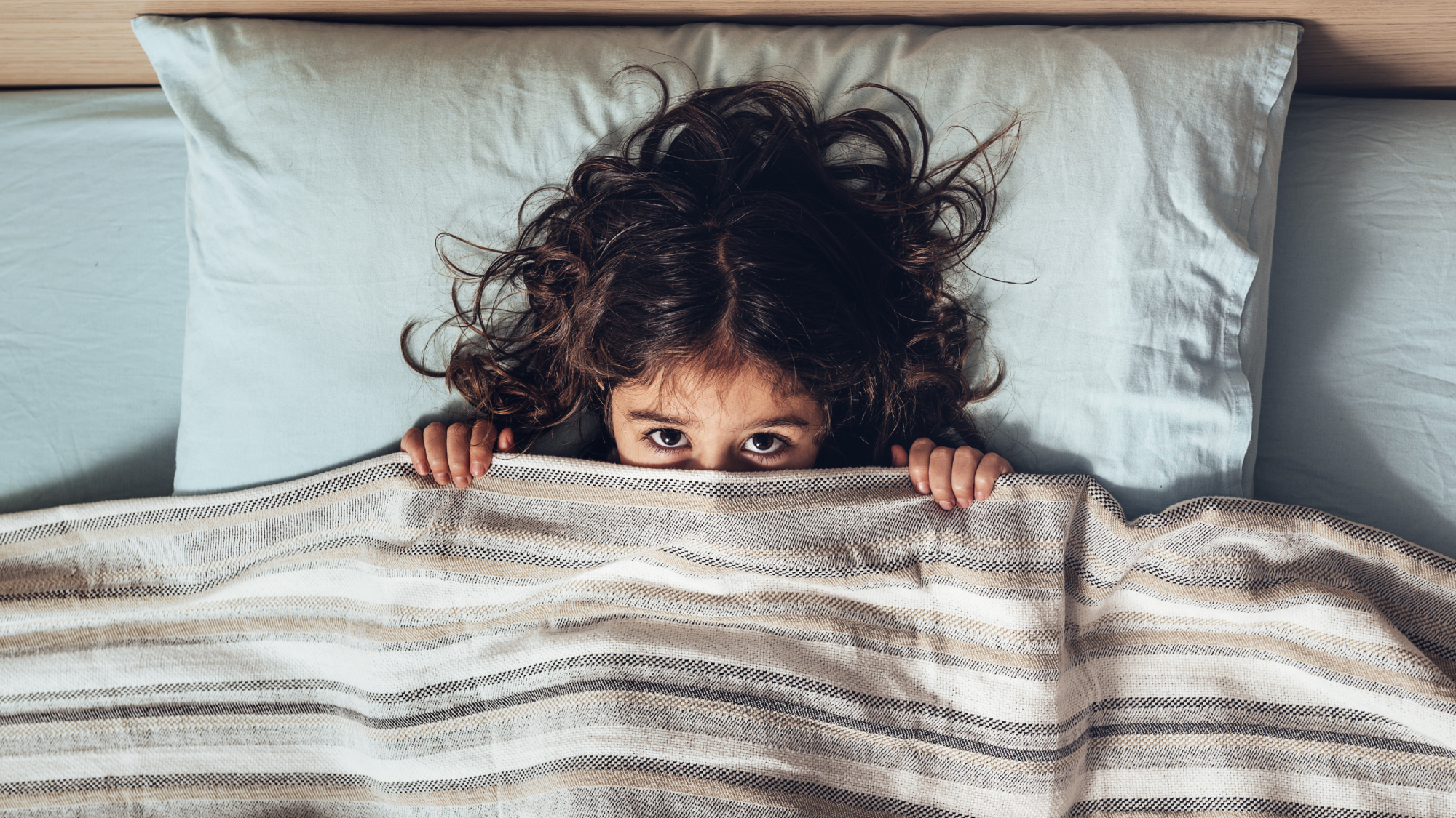 child with dark hair in bed with head on pillow and covers pulled up to nose lookin up into the camera