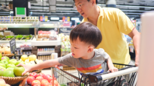 young child in grocery cart seat reaching for produce with a man behind holding on to side of cart