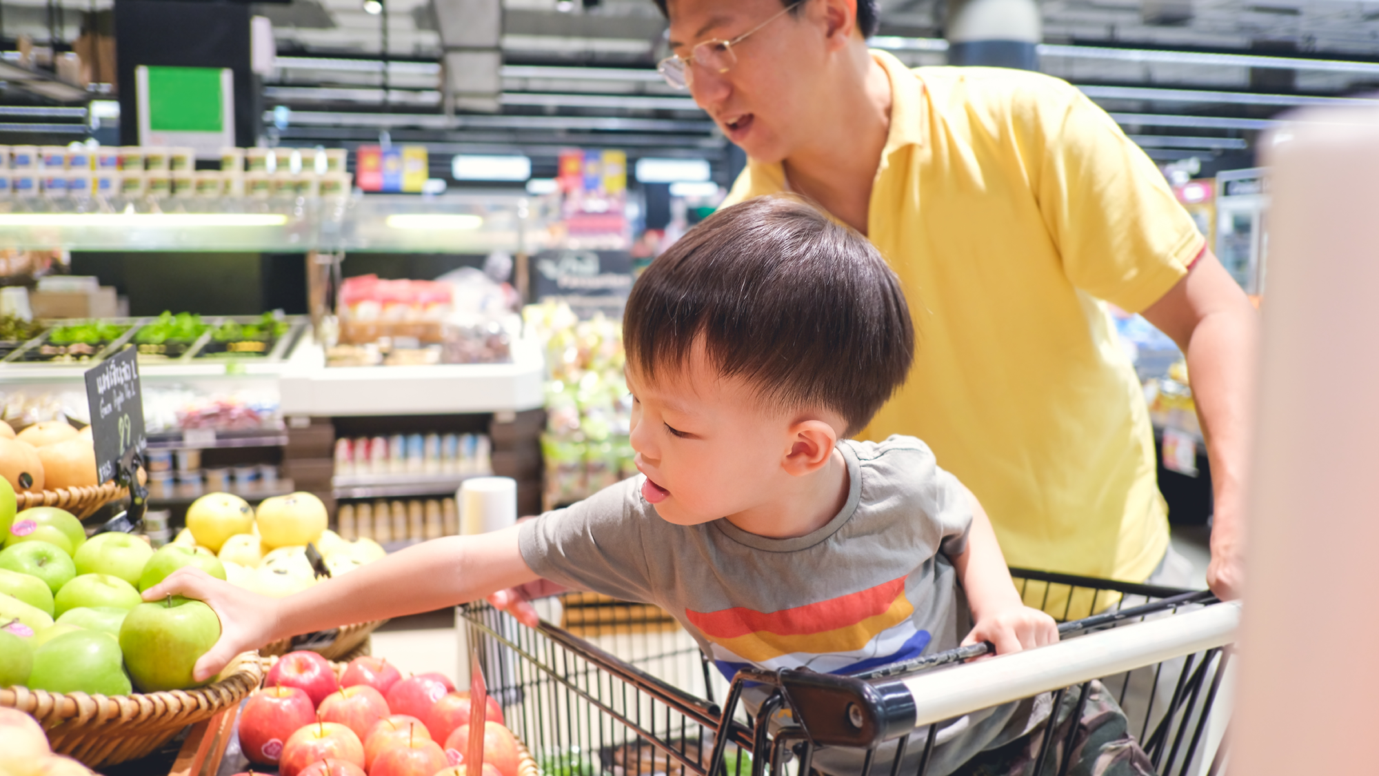 young child in grocery cart seat reaching for produce with a man behind holding on to side of cart