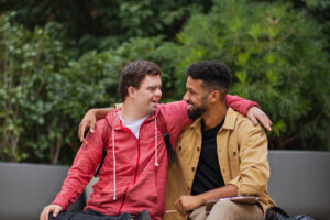 A happy young man with Down syndrome and mentoring friend sitting with arms around outdoors